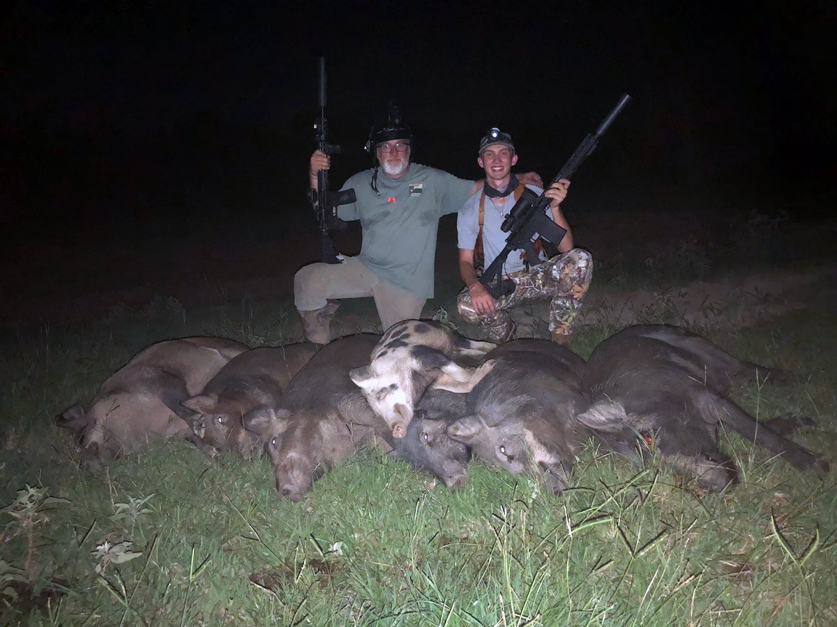 father and sun holding rifles over dead hogs after hunt